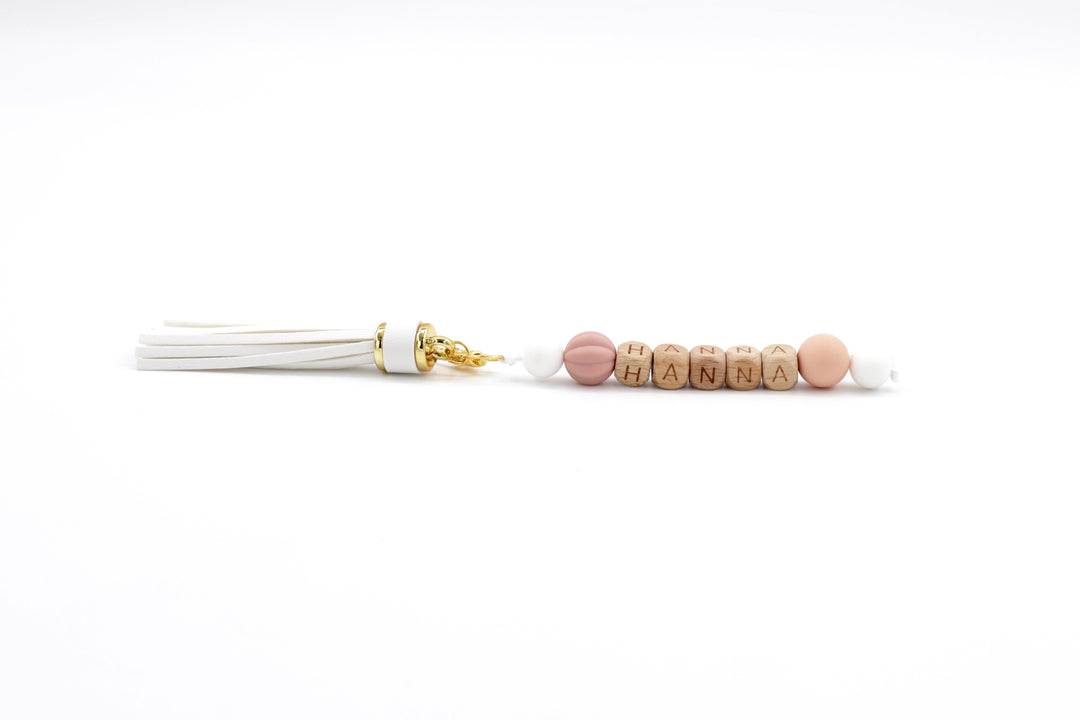 Personalised bag charm with white tassel, wooden letter beads spelling HANNA, and blush-toned silicone accents on white background