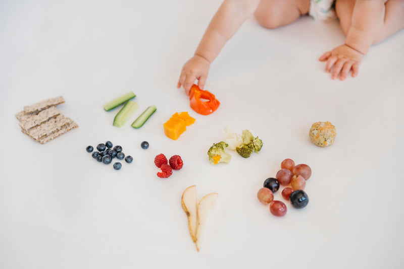 A baby reaches for a red bell pepper slice from a spread of healthy finger foods, including berries, broccoli, and crackers, on a white surface.
