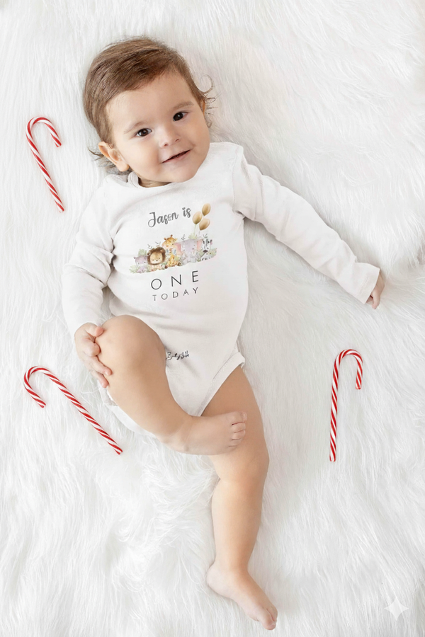 Smiling baby in white onesie reading “Jason is One Today,” lying on fluffy white blanket with candy canes for a Christmas touch.