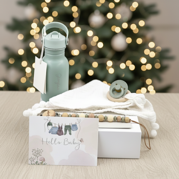 Baby gift set with “Hello Baby” card, personalised water bottle, pacifier, beaded clip, and muslin cloth on table, with Christmas tree in background.
