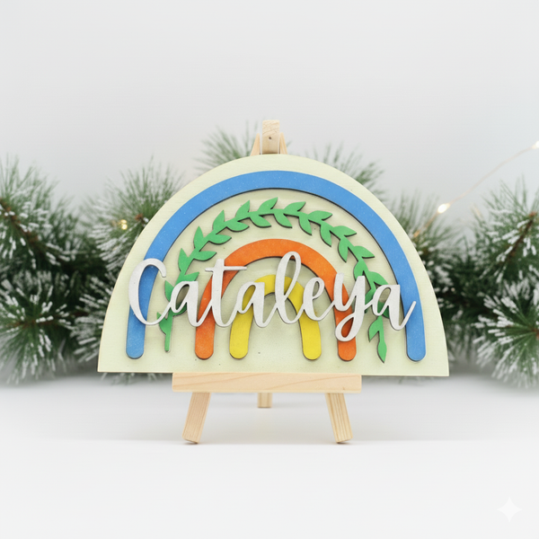 Rainbow-themed wooden sign personalized with the name 'Cataleya'on a mini easel with a blurred christmas garland in the background.