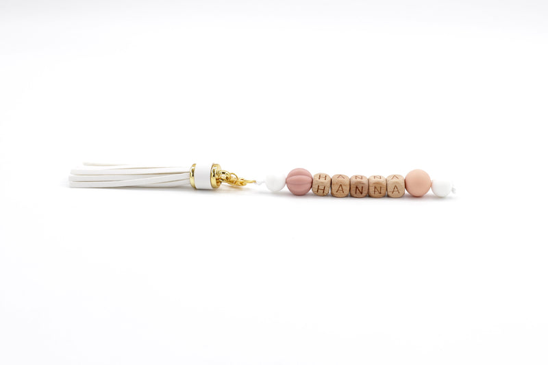 Personalised bag charm with white tassel, wooden letter beads spelling HANNA, and blush-toned silicone accents on white background