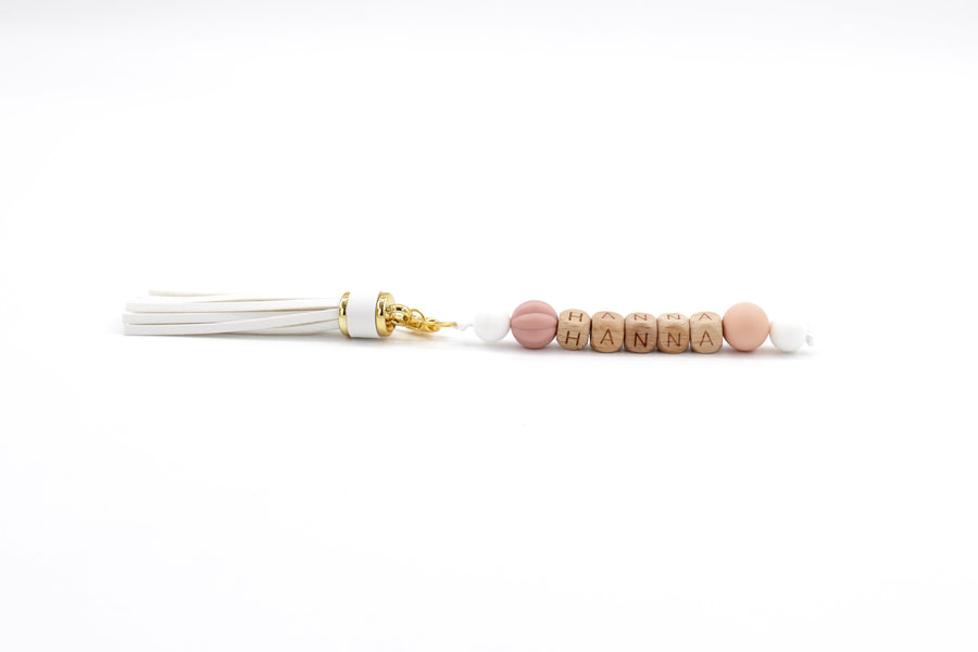 Personalised bag charm with white tassel, wooden letter beads spelling HANNA, and blush-toned silicone accents on white background