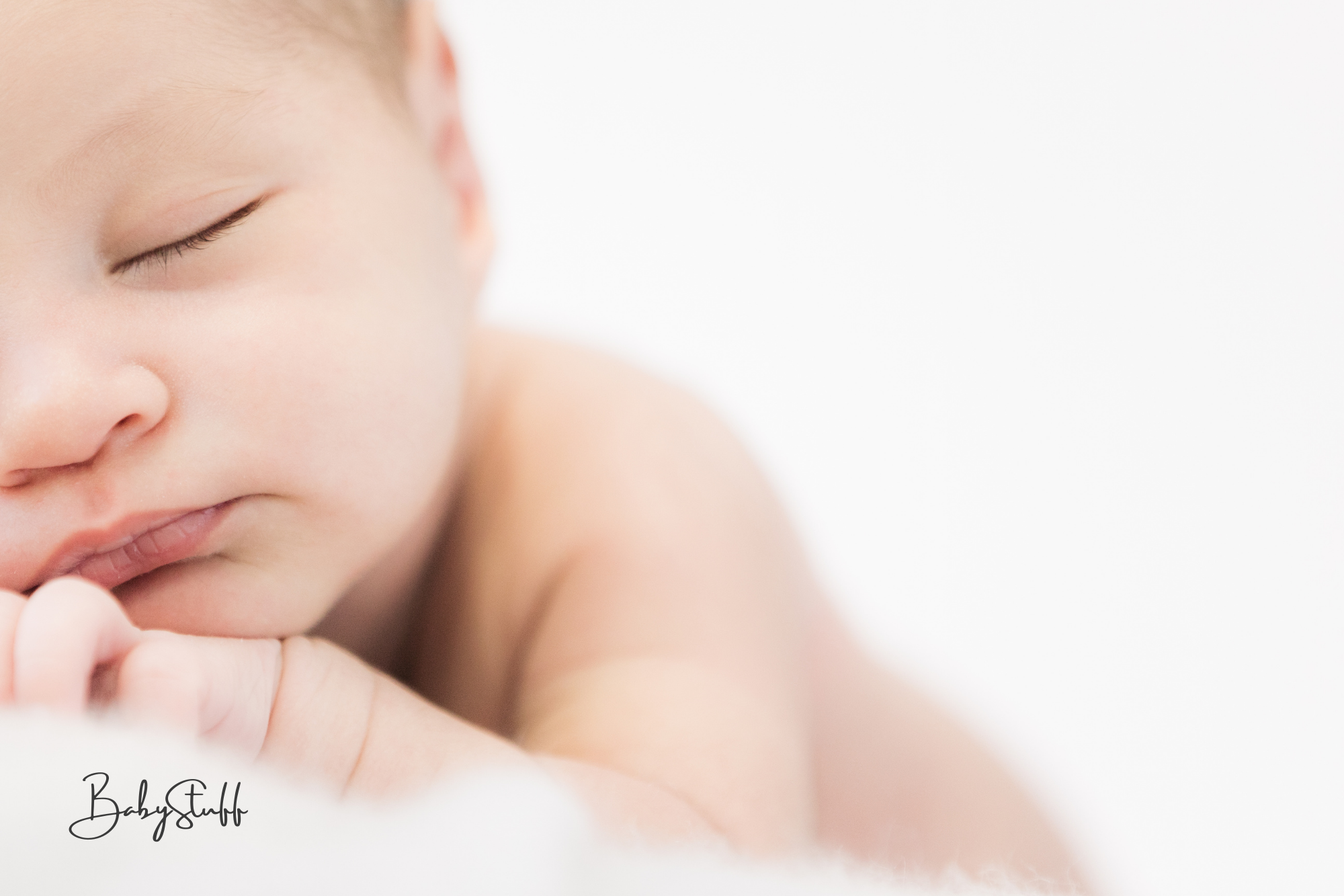 Close-up of a sleeping baby lying on their stomach with eyes closed, face partially visible, and hands tucked under the chin. The background is white and soft-focused.